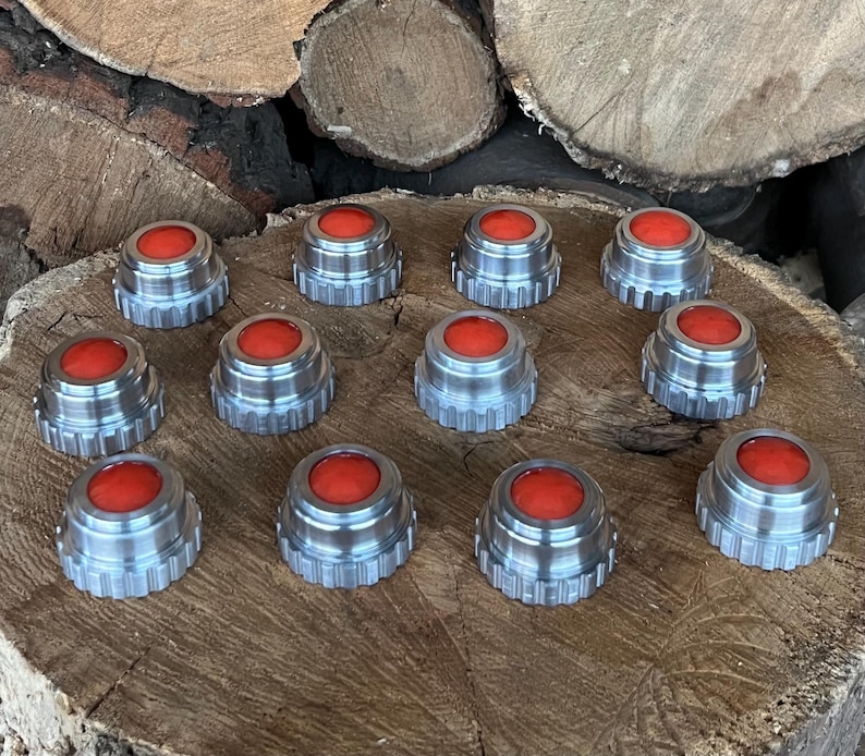 May include: A collection of twelve silver-colored bottle stoppers with a bright red, circular top. The stoppers are arranged on a wooden surface, showcasing their metallic finish and vibrant color contrast. The stoppers are approximately 1 inch in diameter.