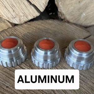 May include: Three aluminum knobs with a red center. The knobs have a ridged edge and a circular top. The word "ALUMINUM" is displayed in a white rectangle below the knobs. The knobs are sitting on a wooden surface.