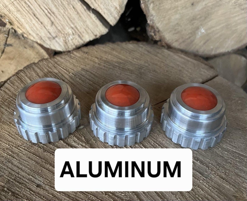 May include: Three aluminum knobs with a red center. The knobs have a ridged edge and a circular top. The word "ALUMINUM" is displayed in a white rectangle below the knobs. The knobs are sitting on a wooden surface.