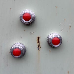 May include: Close-up of three red and silver buttons on a pale green, textured surface. The buttons are circular with a raised, ridged silver edge and a bright red center. The surface shows signs of age and wear.