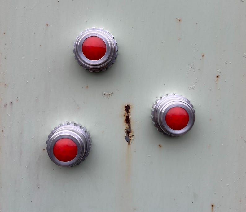 May include: Close-up of three red and silver buttons on a pale green, textured surface. The buttons are circular with a raised, ridged silver edge and a bright red center. The surface shows signs of age and wear.