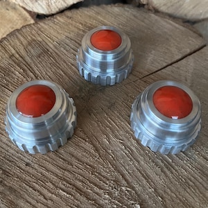 May include: Three metallic bottle stoppers with a red, rounded top. The stoppers have a silver, ridged base and are arranged on a wooden surface. The red tops have a glossy finish.