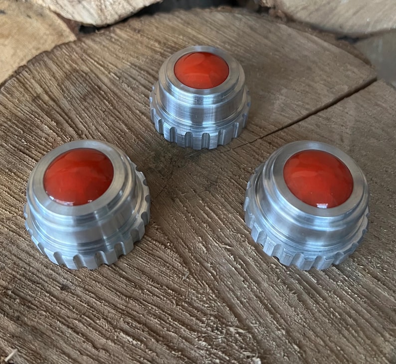 May include: Three metallic bottle stoppers with a red, rounded top. The stoppers have a silver, ridged base and are arranged on a wooden surface. The red tops have a glossy finish.