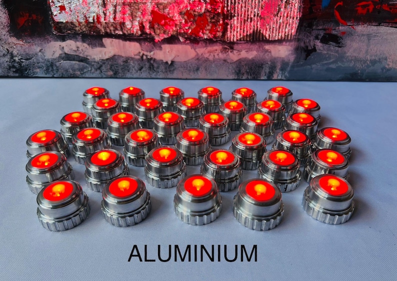 May include: An array of small, cylindrical aluminum objects with a bright red light emanating from the top. The word "ALUMINIUM" is printed in bold, capital letters at the bottom of the image. The background features an abstract painting with red and gray tones.