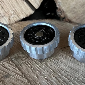 May include: Three silver and black bottle stoppers with a circular design. The stoppers have a ridged, cylindrical base and a black top with a silver center. They are arranged on a wooden surface.
