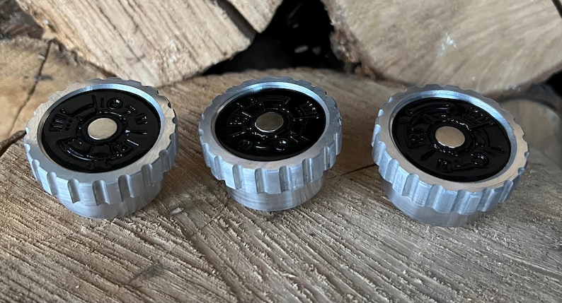 May include: Three silver and black bottle stoppers with a circular design. The stoppers have a ridged, cylindrical base and a black top with a silver center. They are arranged on a wooden surface.