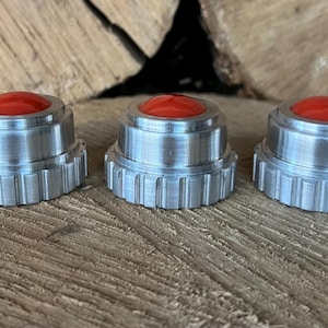 May include: Three silver-colored, machined metal objects with a red, rounded top. The objects have a ridged base and are displayed on a wooden surface. The items appear to be small, possibly decorative or functional.