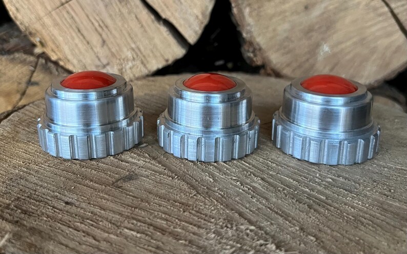 May include: Three silver-colored, machined metal objects with a red, rounded top. The objects have a ridged base and are displayed on a wooden surface. The items appear to be small, possibly decorative or functional.