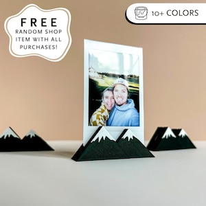 May include: A white framed photo of a couple is displayed in a black and white mountain-shaped photo holder. The holder is made of wood and has white peaks. Additional mountain-shaped holders are visible. The image also includes text that says "FREE RANDOM SHOP ITEM WITH ALL PURCHASES!"