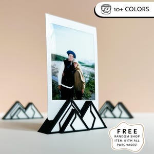 May include: A white photo frame with a black mountain-shaped base holding a photo of two people. The frame has a modern design. The image also includes small mountain-shaped decorations and text that reads "SHIPS IN 48 HRS" and "10+ COLORS".