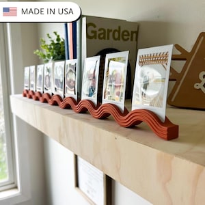 May include: A terracotta-colored photo display stand with a wavy design, holding several small framed photos. The stand is on a wooden shelf, with a book and greenery in the background. The text "MADE IN USA" is in the top left corner.