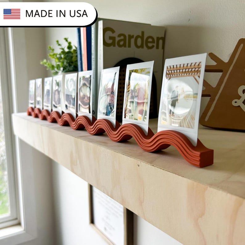May include: A terracotta-colored photo display stand with a wavy design, holding several small framed photos. The stand is on a wooden shelf, with a book and greenery in the background. The text "MADE IN USA" is in the top left corner.
