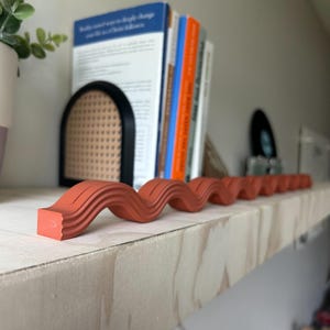 May include: A terracotta-colored, wavy decorative object rests on a light-colored wooden shelf. The object has a series of curved ridges, creating a flowing, serpentine design. Books and a black bookend are in the background, suggesting a home decor theme.
