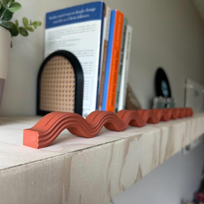 May include: A terracotta-colored, wavy decorative object rests on a light-colored wooden shelf. The object has a series of curved ridges, creating a flowing, serpentine design. Books and a black bookend are in the background, suggesting a home decor theme.