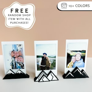May include: Three white photo prints with smiling people, displayed in black and white mountain-shaped holders. The holders have a geometric design. The image also includes text that says "FREE RANDOM SHOP ITEM WITH ALL PURCHASES!" and "10+ COLORS."