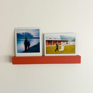 May include: A burnt orange photo ledge displays two framed photographs. One shows a couple by a lake and mountains, the other a wedding scene. The ledge is mounted on a white wall, creating a simple, modern display.