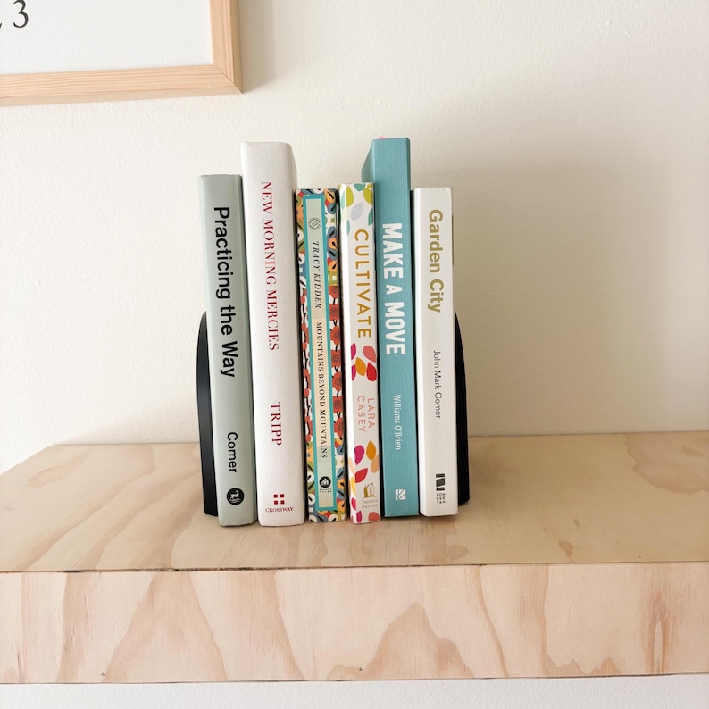 May include: A collection of books with colorful spines, held between two black bookends. Titles include "Practicing the Way," "New Morning Mercies," "Cultivate," and "Make a Move." The books are arranged on a light wood shelf.