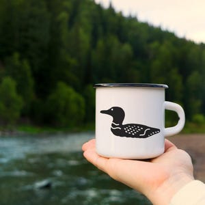 Può includere: Una tazza in smalto bianco con bordo nero e un'illustrazione grafica nera di una strolaga sul lato. La tazza presenta un manico robusto e una finitura liscia e lucida, che mostra una semplice silhouette dell'uccello acquatico di profilo.