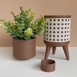 Könnte beinhalten: Ein brauner Pflanztopf mit einer grünen Pflanze, ein brauner Topf und ein brauner Pflanztopf mit weißem Webmuster. Die Artikel sind auf einer weißen Oberfläche mit hellbraunem Hintergrund angeordnet. Artikel für die Inneneinrichtung.