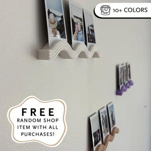 Peut inclure: Étagère à photos blanche en forme de vague, contenant trois photos instantanées. L'étagère est fixée sur un mur blanc, avec des étagères supplémentaires marron et violettes. L'image contient également le texte "FREE RANDOM SHOP ITEM WITH ALL PURCHASES!"