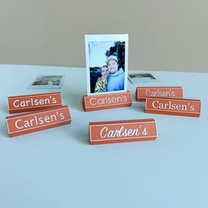 Puede incluir: Varios portafotos naranjas con el nombre "Carlsen's" en escritura blanca. Un portafotos muestra una foto. Los soportes rectangulares tienen un borde blanco y están dispuestos sobre una superficie azul claro.
