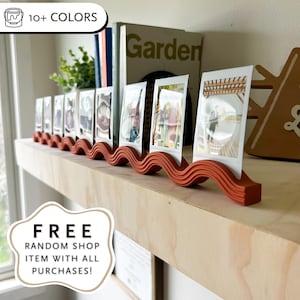 Könnte beinhalten: Ein wellenförmiger, rostfarbener Fotoständer, der mehrere gerahmte Fotos hält. Der Ständer steht auf einem Holzregal, mit Büchern und Grünzeug im Hintergrund. Der Text auf dem Bild lautet "SHIPS IN 48 HRS", "10+ COLORS" und "FREE RANDOM SHOP ITEM WITH ALL PURCHASES!"