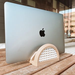 May include: A light brown laptop stand with a woven white wicker design. The stand is shaped like a half-circle and is holding a silver laptop.