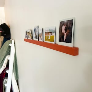 Pode incluir: Uma prateleira flutuante laranja exibe várias fotografias emolduradas contra uma parede branca. As fotos mostram várias cenas, incluindo um casal e paisagens. A prateleira é montada horizontalmente, criando uma exibição moderna.