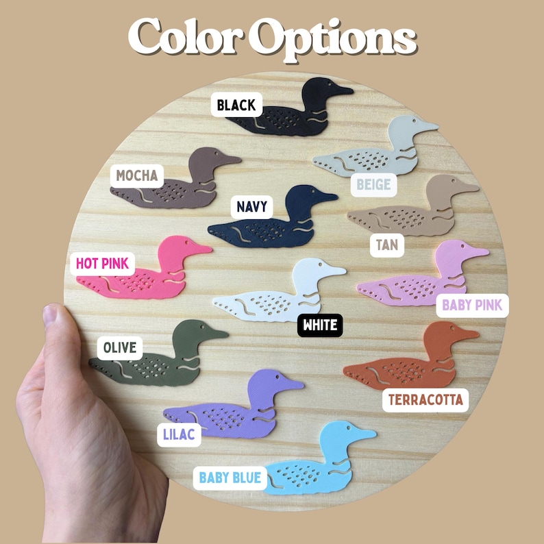 May include: A wooden circle displays various color options for loon-shaped cutouts. Colors include black, mocha, navy, hot pink, olive, lilac, baby blue, beige, tan, baby pink, white, and terracotta. The text "Color Options" is at the top.