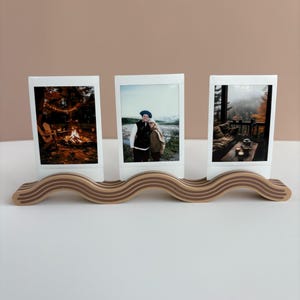 May include: A wooden photo display stand with a wavy design, holding three instant photos. The photos depict outdoor scenes, including a campfire, a couple, and a cabin. The stand is made of light brown wood.