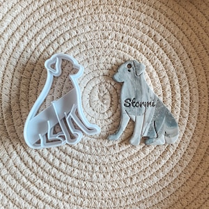 May include: Two white plastic dog cookie cutters, one with the name "Stormi" engraved on it. The cutters are on a woven brown background.