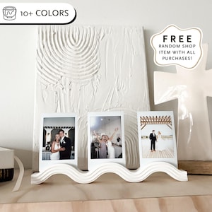 May include: White, wavy photo display holding three framed wedding photos. A textured white art piece and a white cross-shaped decoration are in the background. Text on the image says "Get Your Photos" and "10+ Colors."