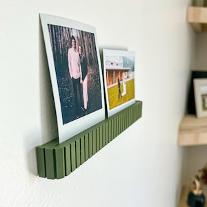 May include: A green, grooved photo shelf displays two framed photographs. The shelf is mounted on a white wall, showcasing the photos. The photos appear to be of a couple in a forest and a wedding.