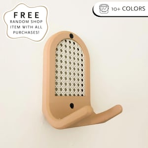 Könnte beinhalten: Ein wandmontierter Kleiderhaken in Hellbraun. Der Haken hat ein gebogenes Design mit einem Einsatz aus gewebtem Rohrgeflecht und einem einzelnen Haken. Der Haken wird mit zwei schwarzen Schrauben an der Wand befestigt. Ein dekorativer und funktionaler Artikel.