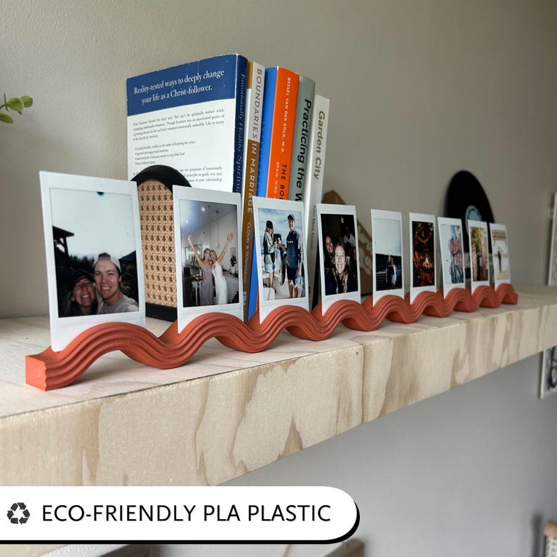 May include: A terracotta-colored photo display with a wavy design, holding several instant photos. Books are placed behind the photos. The text "ECO-FRIENDLY PLA PLASTIC" is at the bottom.
