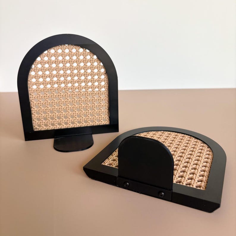 May include: Two black metal arched bookends with woven cane inserts. One bookend is upright, the other is lying flat. The bookends have a modern design and are set against a neutral background.