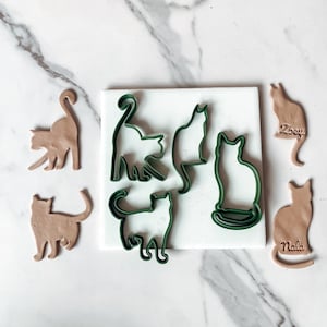 May include: A set of five green cat-shaped cookie cutters on a white surface, with five brown cat-shaped cookie dough cutouts. The cookie cutters are arranged in a circle, and the cookie dough cutouts are arranged in a line. The cookie cutters are labeled "Zoey" and "Nala".
