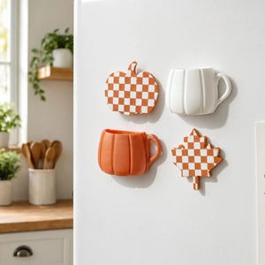 May include: Four decorative magnets attached to a white surface. The set includes a flat orange and white chequered pumpkin, a white pumpkin-shaped mug, a solid orange pumpkin-shaped mug, and a flat orange and white chequered maple leaf.