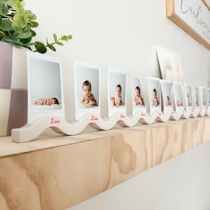 Peut inclure: Étagère en bois avec une série de photos de bébé dans des cadres blancs. Les photos sont disposées par ordre chronologique, avec les mois étiquetés en rose. Une pièce décorative en bois avec un design ondulé maintient les cadres.