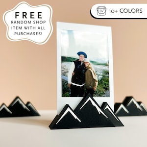 May include: Black mountain-shaped photo holder with white accents, holding a photo. The photo holder is designed to resemble a mountain range. The image also includes text that says "FREE RANDOM SHOP ITEM WITH ALL PURCHASES!" and "10+ COLORS."