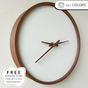 Op de afbeelding: Een moderne wandklok met een minimalistisch ontwerp. Het heeft een witte wijzerplaat, een bruin cirkelvormig frame en bruine wijzers. De klok is aan een witte muur bevestigd. De afbeelding bevat ook de tekst "FREE RANDOM SHOP ITEM WITH ALL PURCHASES!"