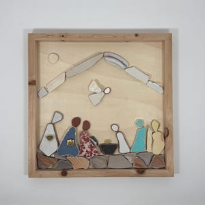 May include: A framed mosaic artwork depicting a nativity scene. The artwork features figures and elements crafted from various materials, including stone and ceramic. The color palette includes beige, brown, blue, and red, with a wooden frame.
