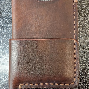 Può includere: Un portafoglio in pelle marrone con fessura per carte. Il portafoglio è realizzato in pelle marrone scuro e presenta cuciture visibili sui bordi. Il design è semplice e funzionale, adatto all'uso quotidiano. Misura circa 10 cm di altezza.