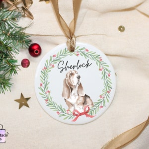 May include: White ceramic Christmas bauble featuring a watercolour illustration of a basset hound dog wearing a red bow. The dog is surrounded by a green wreath with red berries. The bauble is personalised with the name "Sherlock".