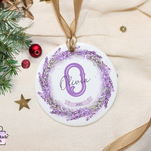 May include: A white ceramic bauble with a lavender wreath and the letter "O" in purple with the name "Olivia" and the year "2023" written on it.