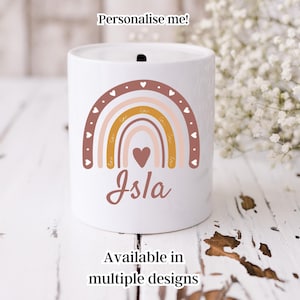 Könnte beinhalten: Weiße Keramik-Sparbüchse mit einem Regenbogen-Design in den Farben Rosa, Braun und Gelb. Der Regenbogen hat ein Herz in der Mitte und der Name "Isla" steht darunter. Der Text "Personalisieren Sie mich!" befindet sich oben im Bild.