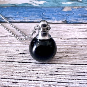 Könnte beinhalten: Eine schwarze, runde Anhängerhalskette mit einer silberfarbenen Kette. Der Anhänger hat einen glänzenden, kugelförmigen schwarzen Körper und eine silberfarbene Kappe. Die Halskette wird auf einer hellen Holzoberfläche mit blauen Akzenten präsentiert.