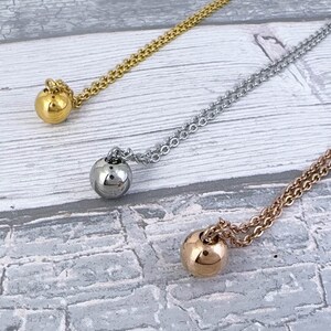 May include: Three necklaces with ball pendants in gold, silver, and rose gold. Each necklace features a delicate chain and a smooth, spherical pendant. The necklaces are displayed on a light gray and white wooden surface.