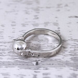 May include: A silver ring with a cat design. The ring features a rounded cat head and a detailed band. The inscription "S925" is visible on the band. The ring is set against a light-colored, textured background.