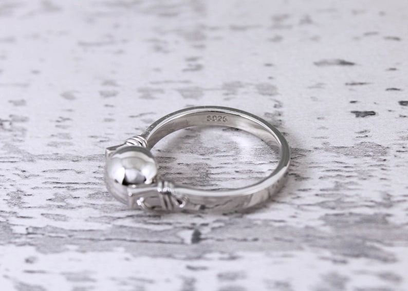 May include: A silver ring with a cat design. The ring features a rounded cat head and a detailed band. The inscription "S925" is visible on the band. The ring is set against a light-colored, textured background.